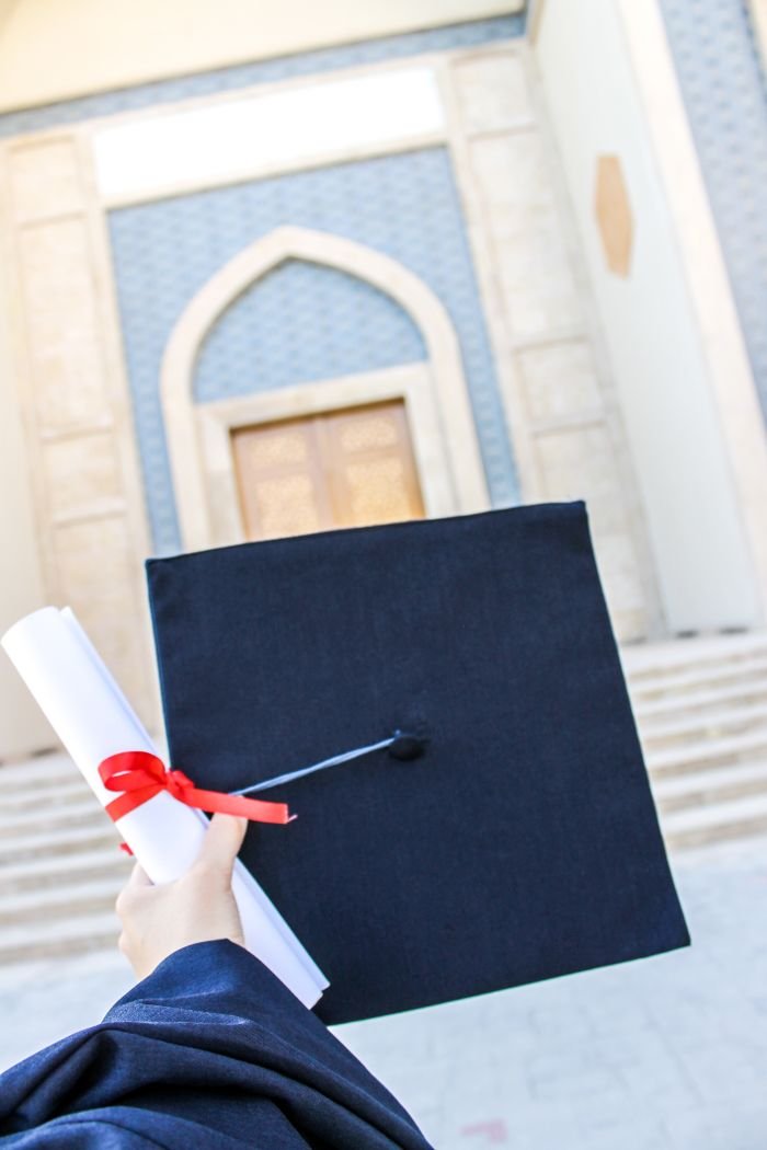 A hand holding a graduation hat and diploma in front of a university building.